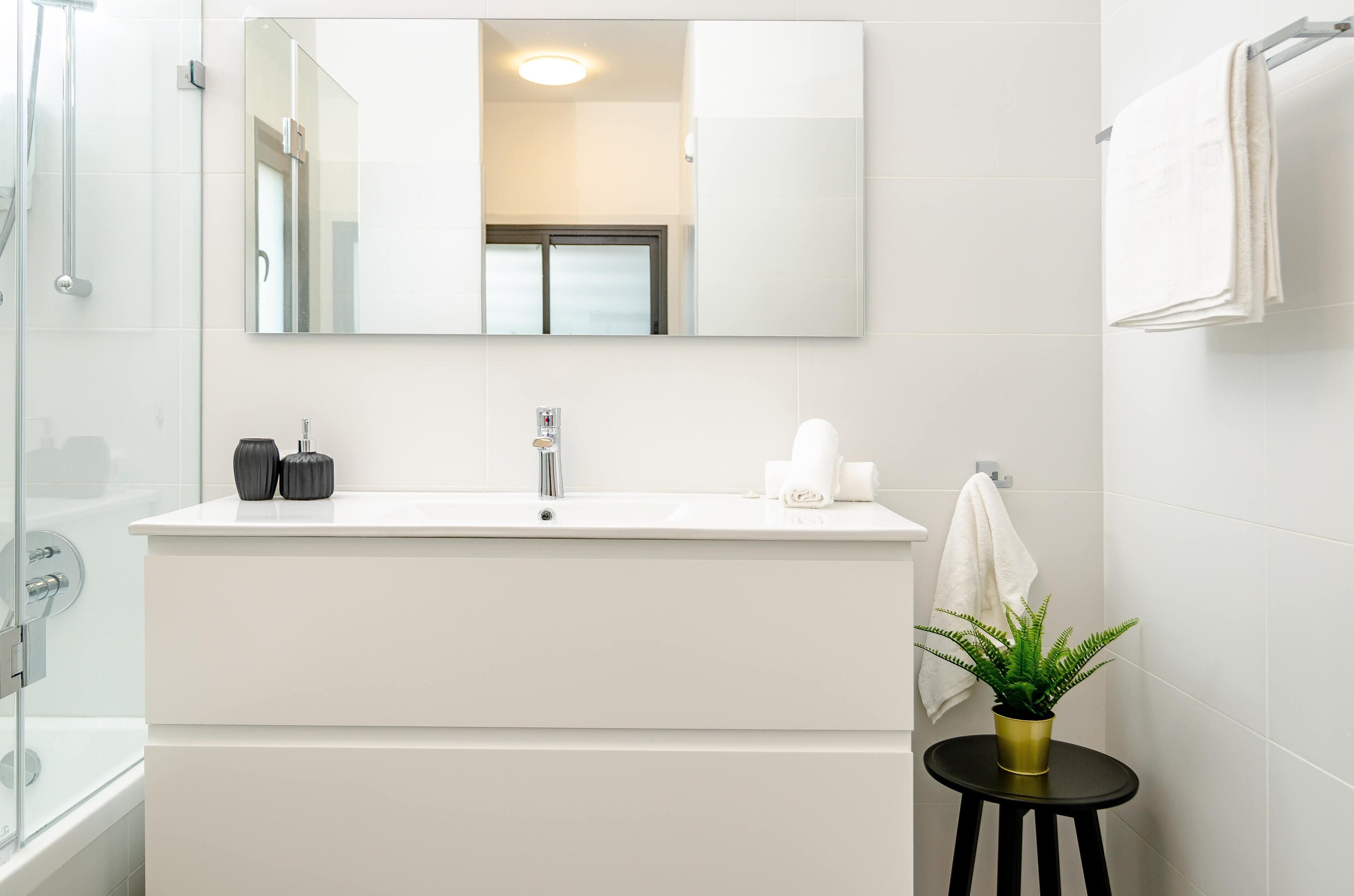 A modern bathroom with a large mirror, a white sink, a shower, and a small stool with a plant.