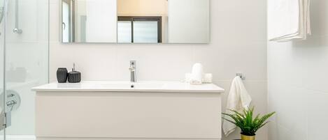 A modern bathroom with a large mirror, a white sink, a shower, and a small stool with a plant.