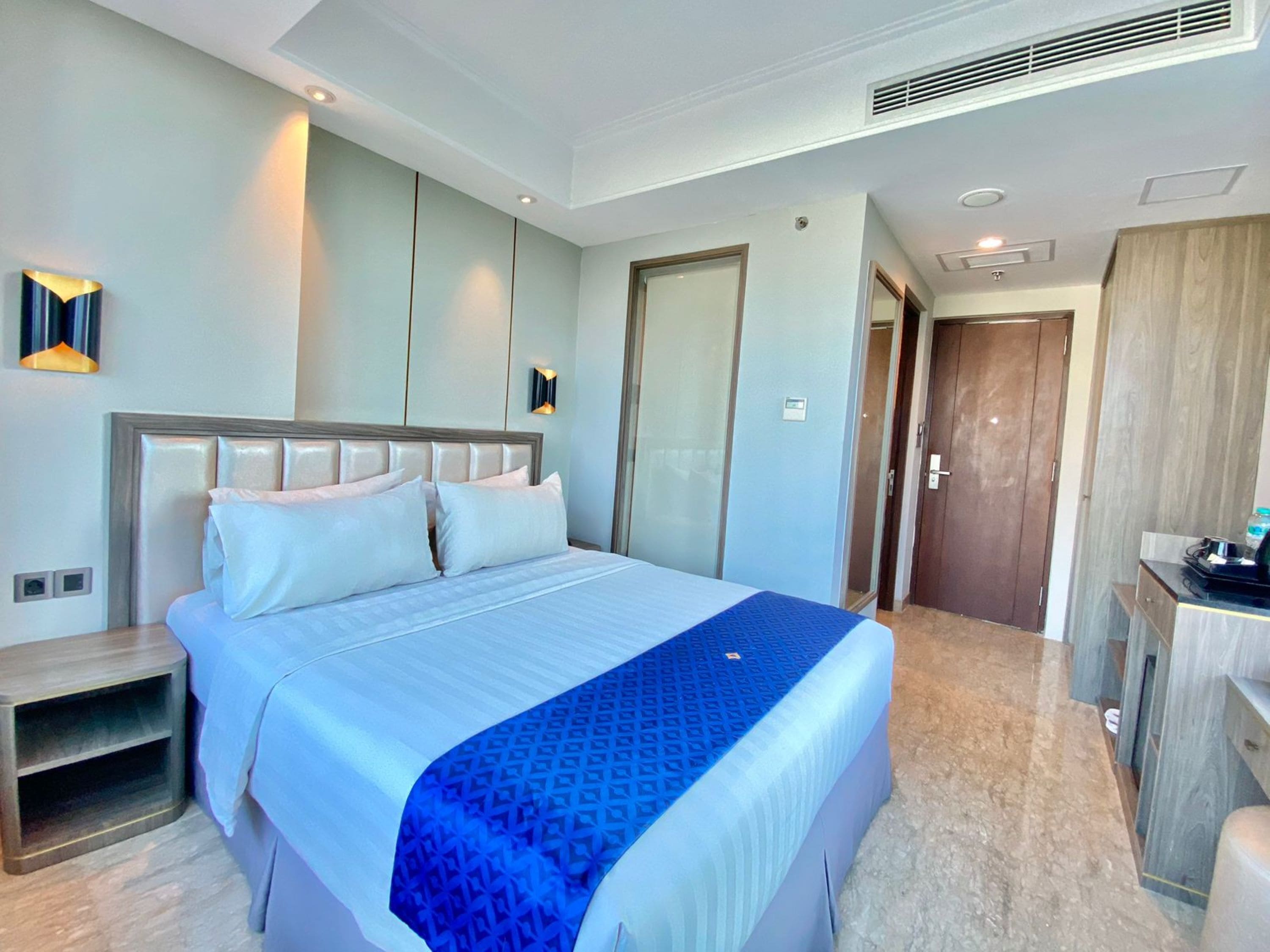 A modern hotel room with a large bed, a bedside table, a wardrobe, and a desk with a chair.