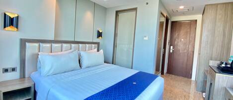 A modern hotel room with a large bed, a bedside table, a wardrobe, and a desk with a chair.