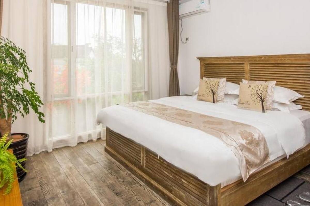 Een slaapkamer met een groot bed, een houten hoofdbord, een potplant en een raam met dunne gordijnen.