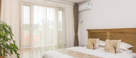 A bedroom with a large bed, a wooden headboard, a potted plant, and a window with sheer curtains.