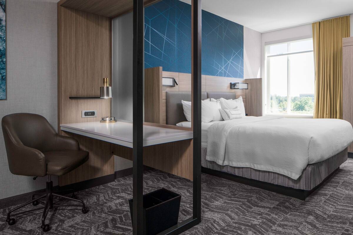 A hotel room with a bed, a desk, a chair, a mirror, and a window with curtains.