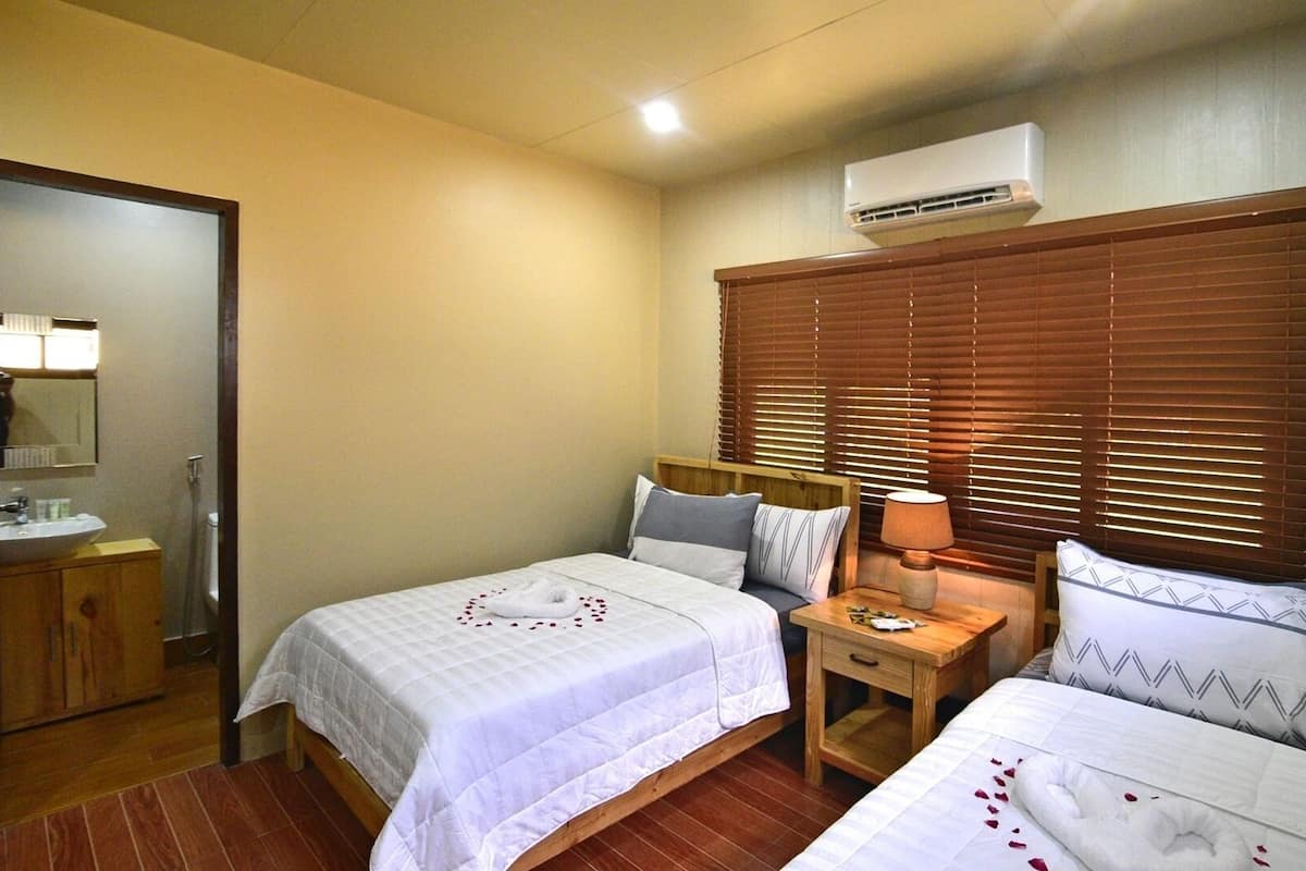 Een hotelkamer met twee bedden, een houten hoofdbord, een nachtkastje met een lamp en een airconditioner boven het raam.