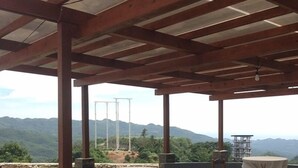 Ein Terrassenbereich im Freien mit Holzbalken, runden Tischen und Stühlen, umgeben von angelegten Gärten und einer Bergkulisse.