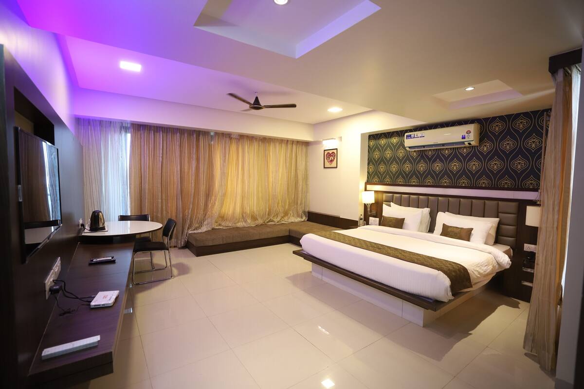 A hotel room with a large bed, a desk with a computer, a TV, and an air conditioning unit.