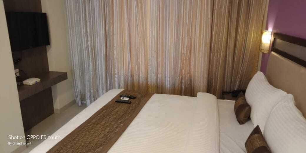 A hotel room with a bed, a bedside table with a lamp, a TV on a shelf, and a curtain.