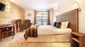 A hotel room with a wooden floor, a bed with a fur throw, a desk with a chair, a TV mounted on the wall, and a balcony with curtains.