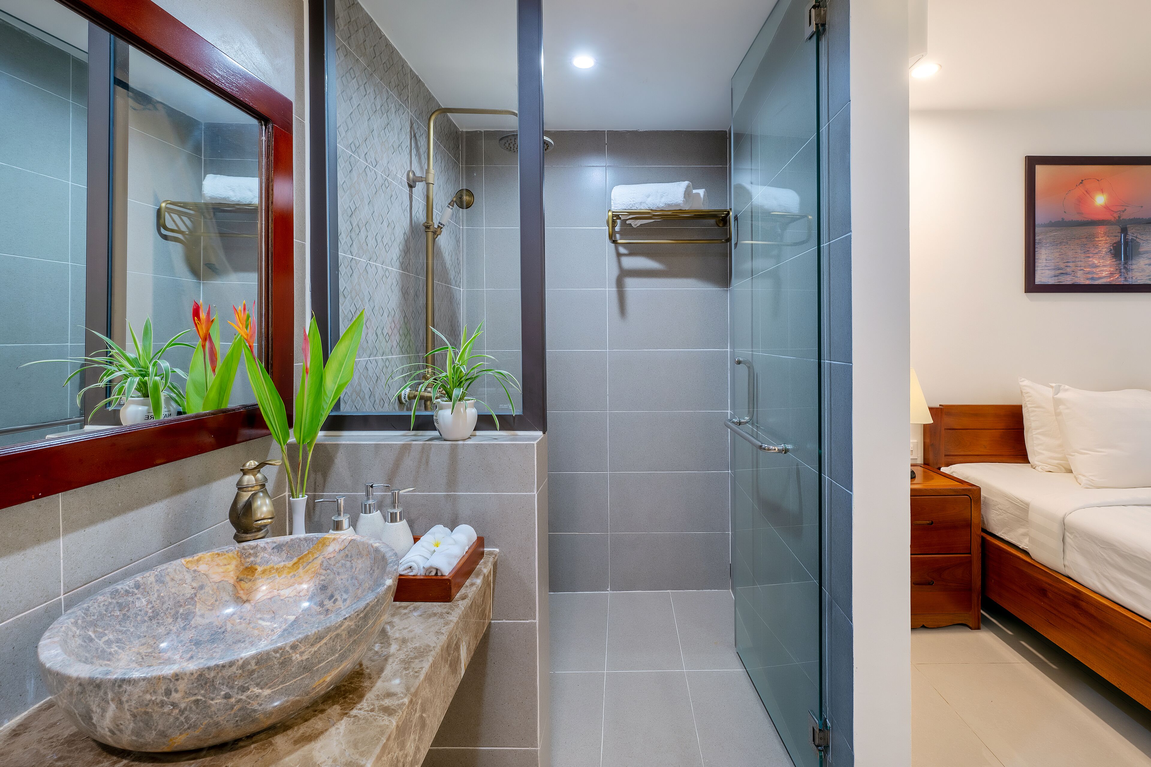 A modern bathroom with a marble sink, two potted plants, a glass shower enclosure, and a bed with white bedding.