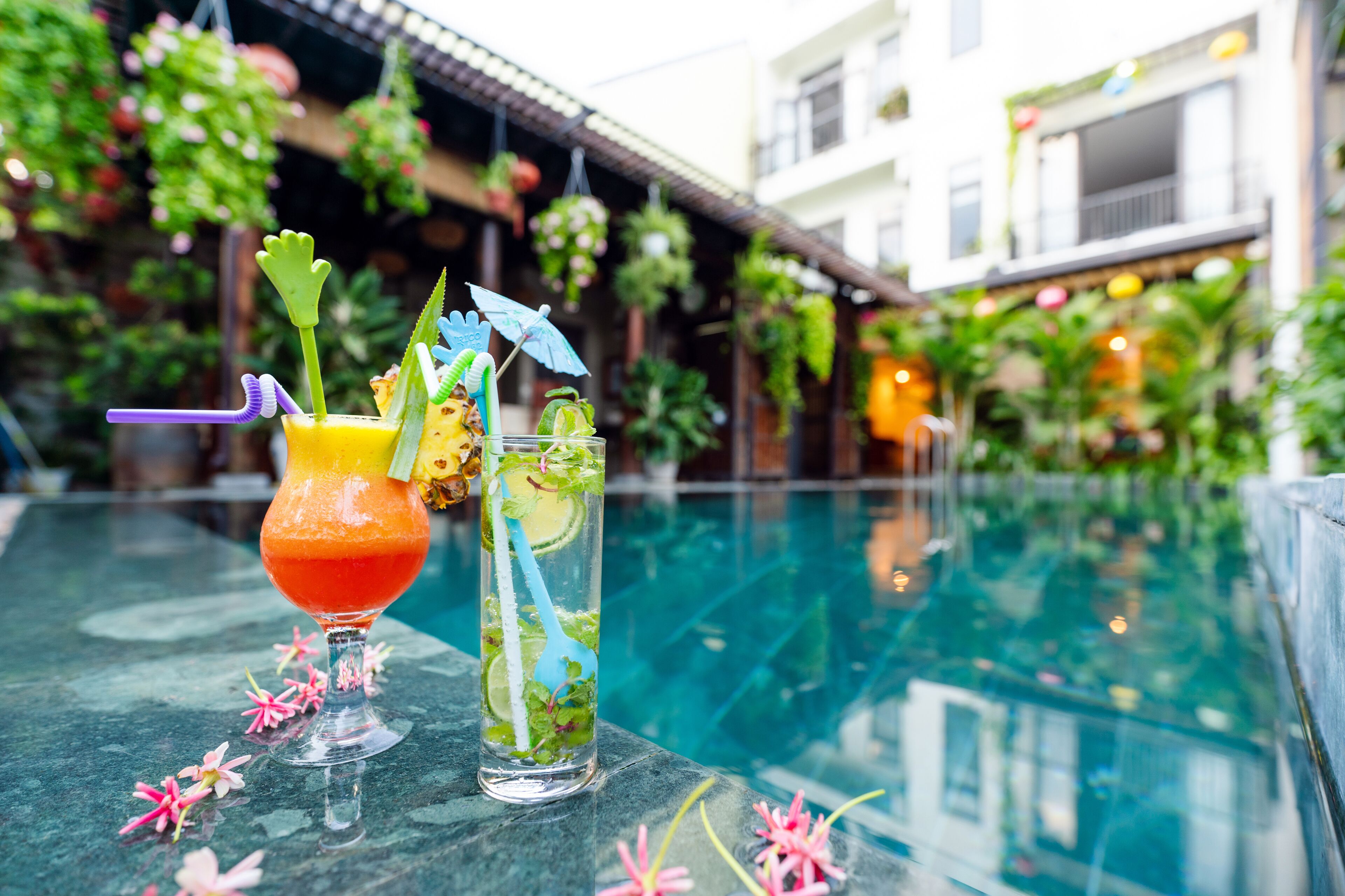Two cocktails on a table near a pool with a view of a building and hanging plants.
