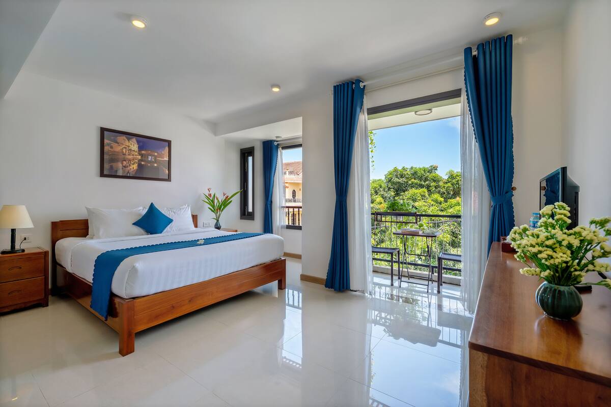 Habitación de hotel con una cama de madera, ropa de cama blanca, un balcón con cortinas azules y vistas a zonas verdes.