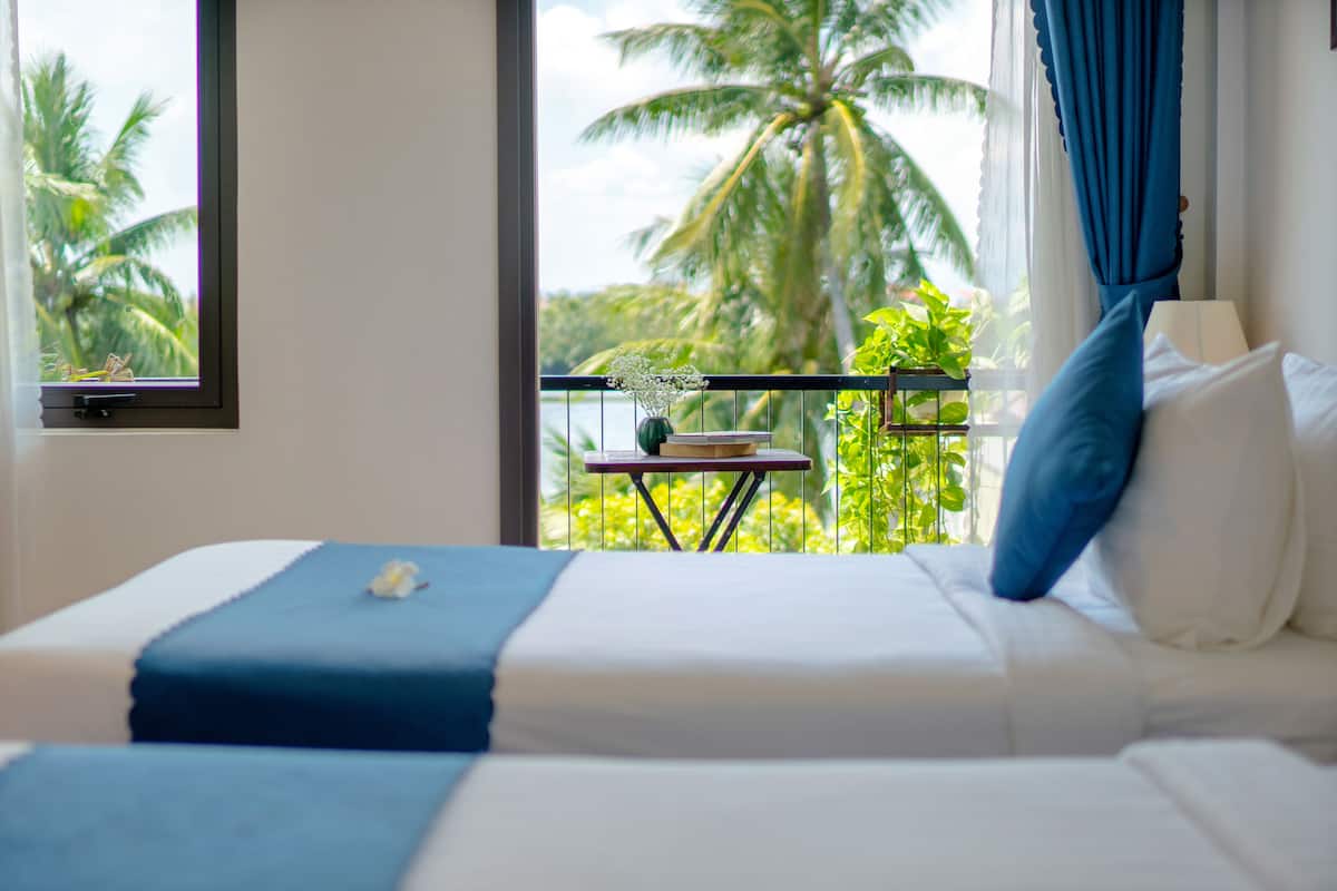 Habitación de hotel con cama, una mesita con una planta y vista a palmeras a través de una ventana.