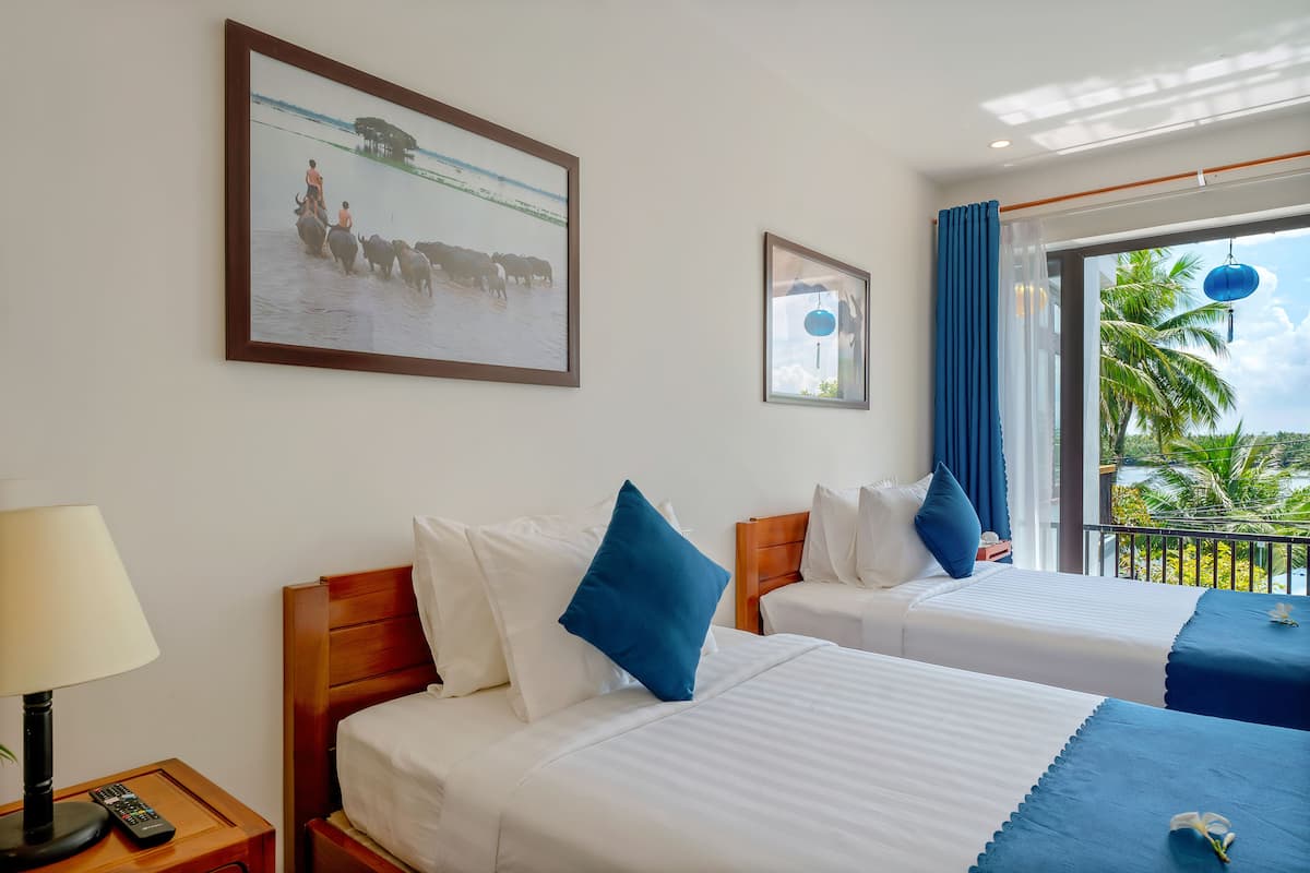 Habitación de hotel con dos camas, una mesita de noche con lámpara y control remoto, y un ventanal con vistas a palmeras.