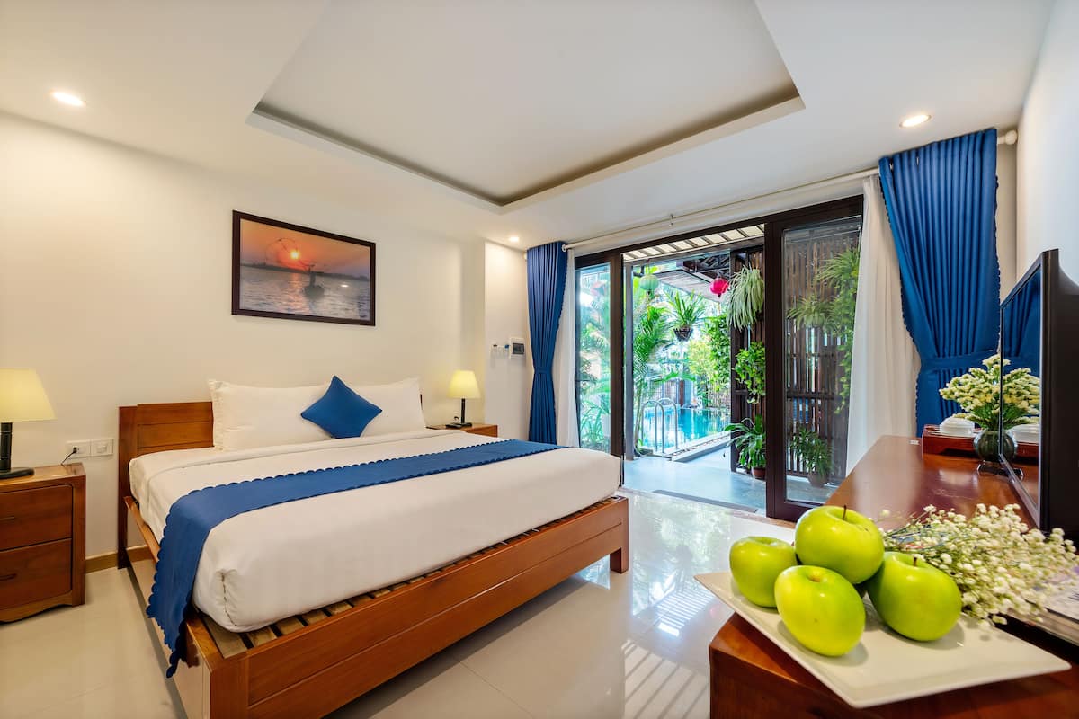Una habitación de hotel con cama, mesita de noche, una lámpara, ventana con cortinas y vistas a una piscina y follaje exterior.