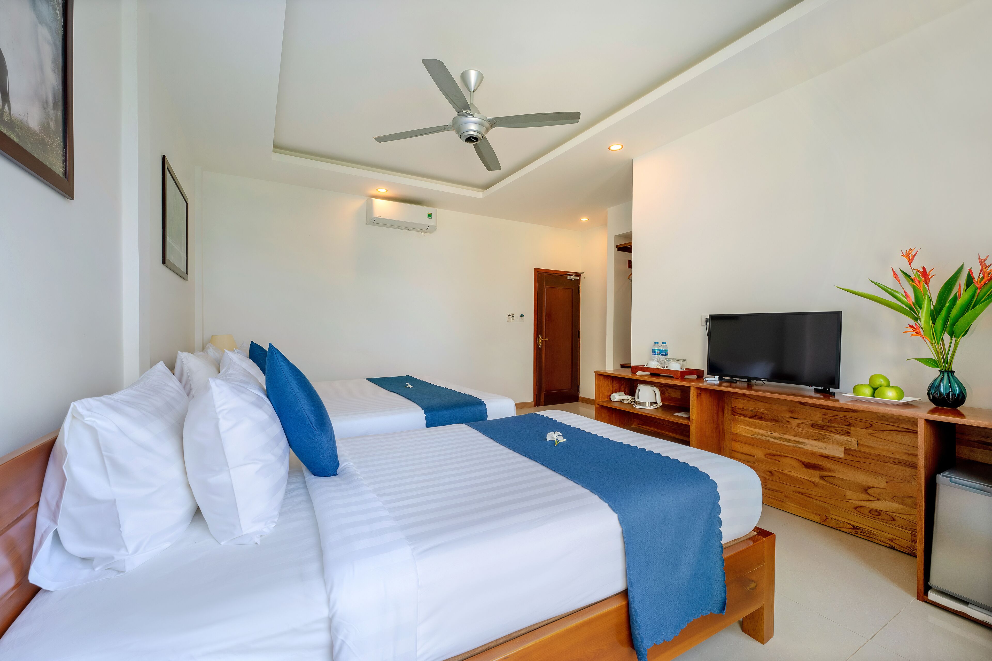 Habitación de hotel con dos camas, ventilador de techo, televisión, cómoda de madera y un refrigerador pequeño.
