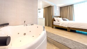 Une chambre d’hôtel moderne avec une grande baignoire, un lit avec du linge de maison blanc et des rideaux de couleur beige.