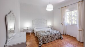 Une chambre à coucher comprenant un lit, des tables de chevet, une commode, un miroir, une lampe suspendue et une fenêtre avec des rideaux.