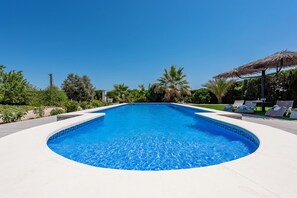 A large, clear blue swimming pool with a curved edge, surrounded by a white deck. There are lounge chairs and a thatched umbrella on the right side. - Casa Manuel, nice holiday home in Seville Countryside (Puebla De Cazalla, La)