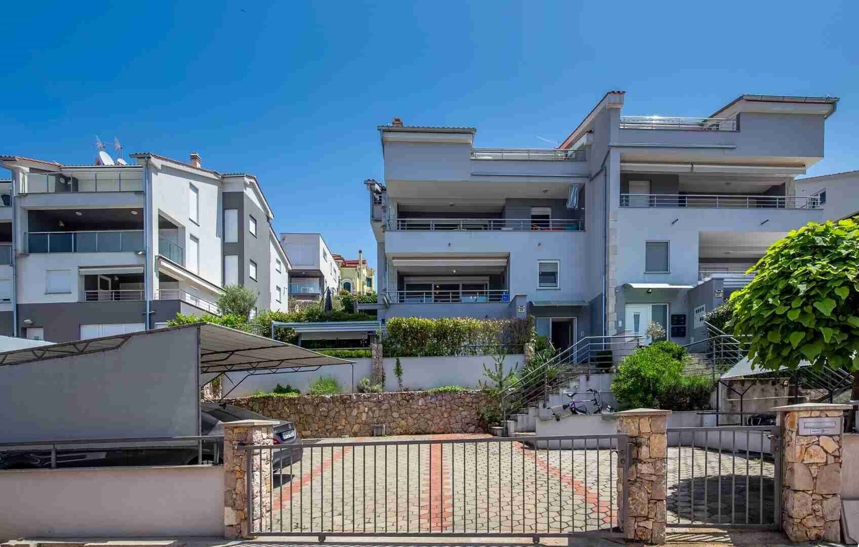 Un edificio residencial moderno de varios pisos, con balcones, entrada con portón y un muro de piedra.