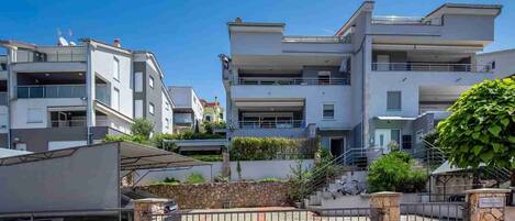Un edificio residencial moderno de varios pisos, con balcones, entrada con portón y un muro de piedra.