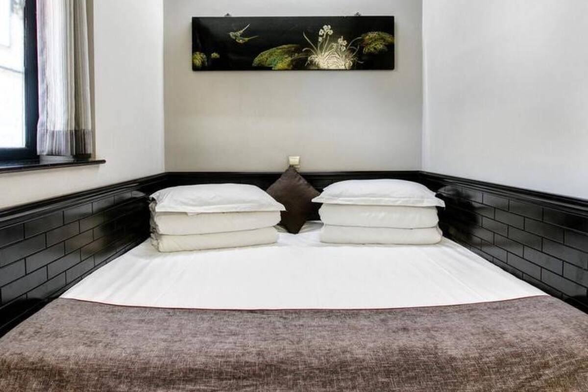 Un lit bien fait, avec des draps blancs et deux oreillers, une couverture en laine marron foncé et une œuvre d’art accrochée au mur représentant la nature.