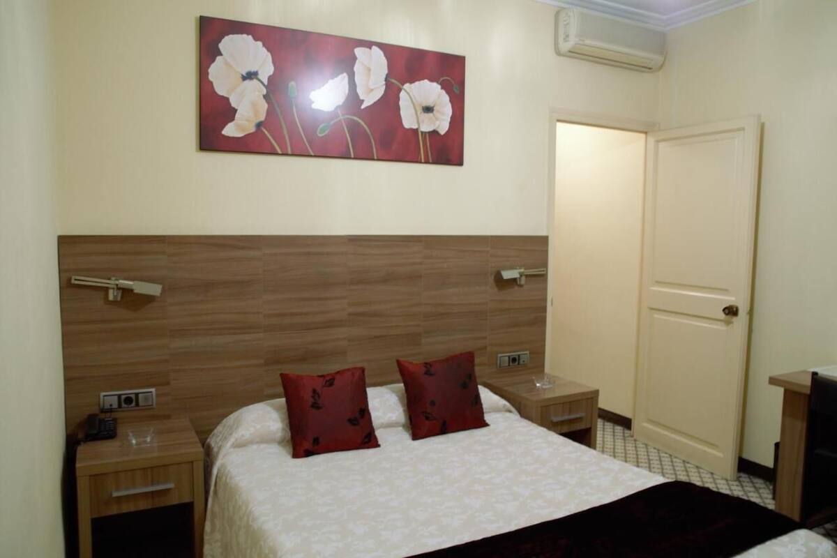 A hotel room with a wooden headboard, two red pillows, a bedside table with a lamp, and a painting of white flowers on the wall.