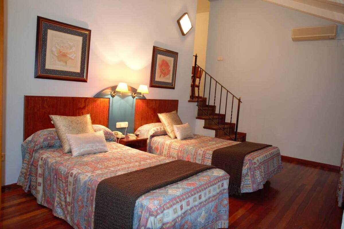 A hotel room with two beds, a wooden headboard, a nightstand, a lamp, and a staircase.