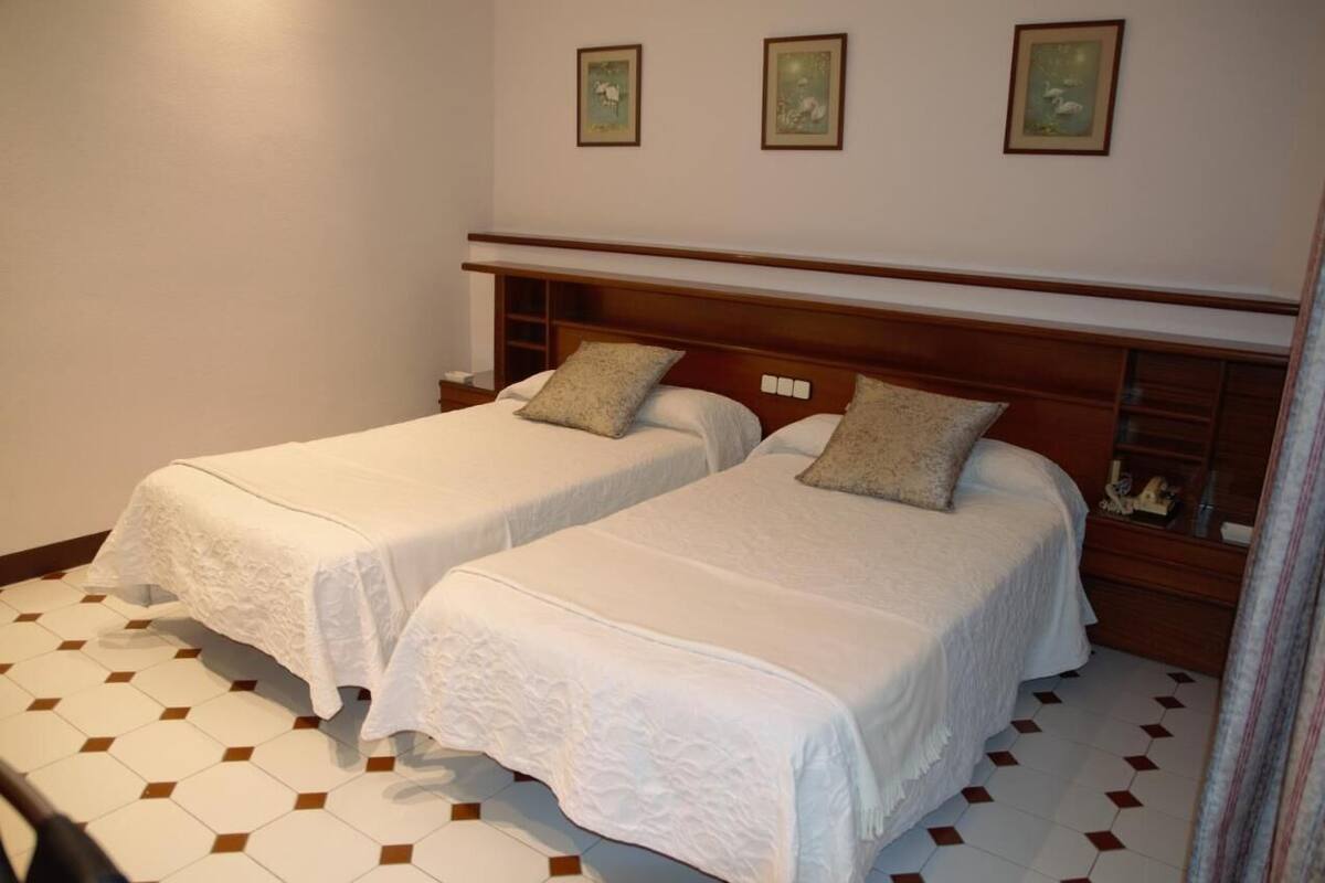 A hotel room with two single beds, a wooden headboard, a small shelf with decorative items, and a tiled floor.