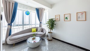 A modern living room with a grey sofa, a glass coffee table, a potted plant, and a view through large windows with blue curtains. - Floral Hotel Jiu Qi Yin Ju Dunhuang (Jiuquan)