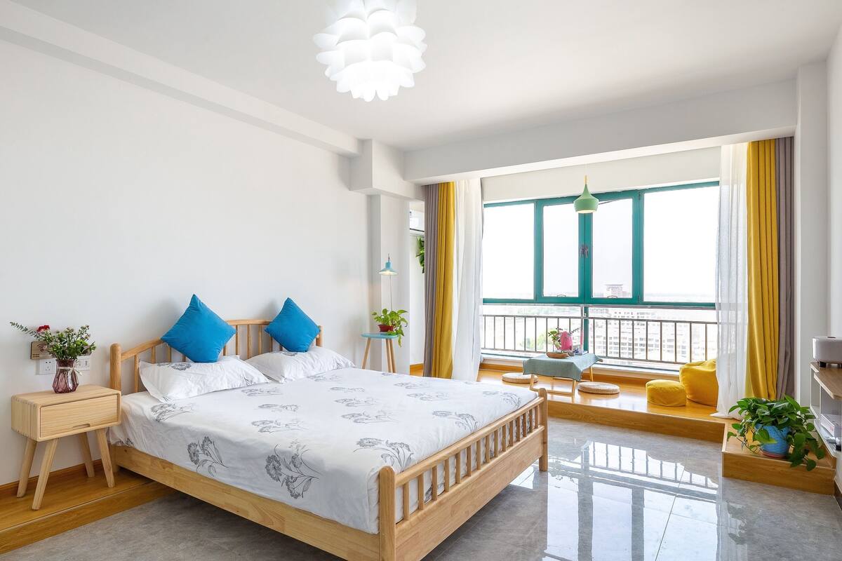 Ein modernes Schlafzimmer mit einem Holzbett, einem Nachttisch, einem kleinen Tisch mit einer Pflanze und einem Balkon mit gelben Vorhängen.