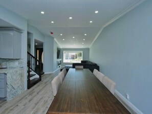 A spacious living room with a long wooden dining table, a black sofa, and a kitchen area with marble countertops.