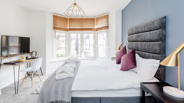 A modern hotel room with a large bed, a desk with a chair, a television, and a view of the city through large windows. - Westland House Guest house (Watford)