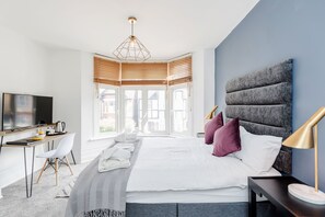 A modern hotel room with a large bed, a desk with a chair, a television, and a view of the city through large windows. - Westland House Guest house (Watford)