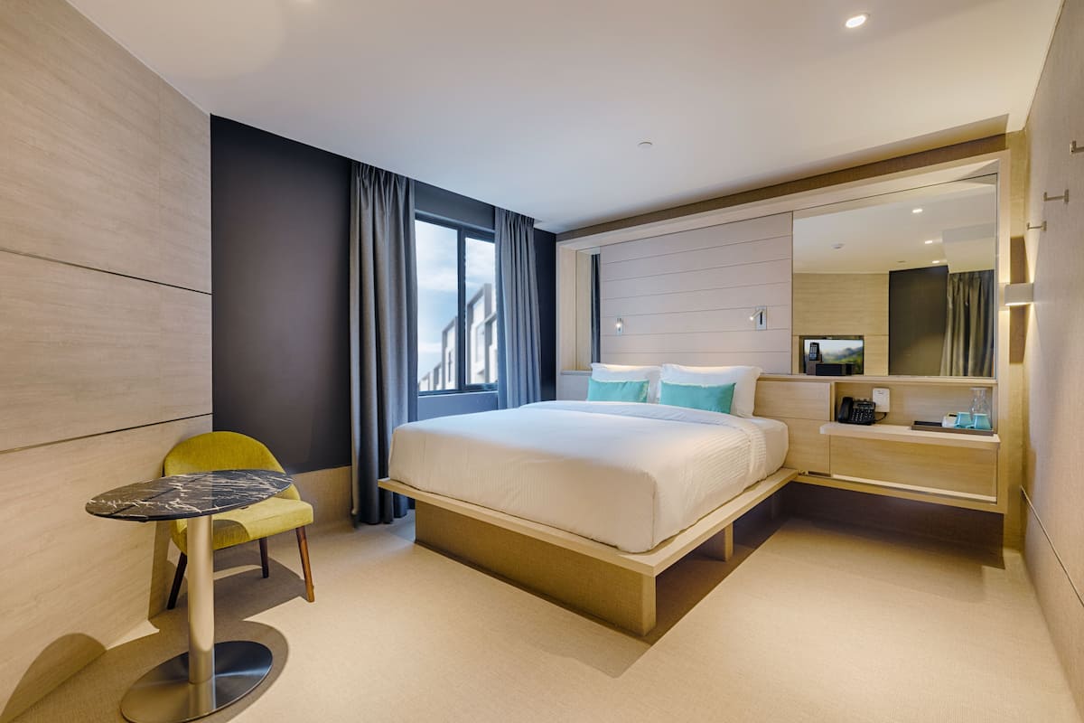 A modern hotel room with a large bed, a small table, a chair, and a built-in desk with a mirror.