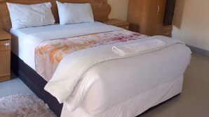A neatly made bed with white bedding and a floral-patterned blanket, a wooden wardrobe, and a fluffy rug on a tiled floor. - Kamuboola Lodge (Choma)