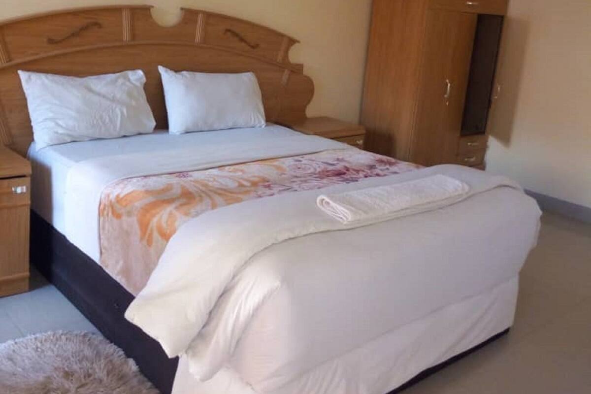 A neatly made bed with white bedding and a floral-patterned blanket, a wooden wardrobe, and a fluffy rug on a tiled floor.