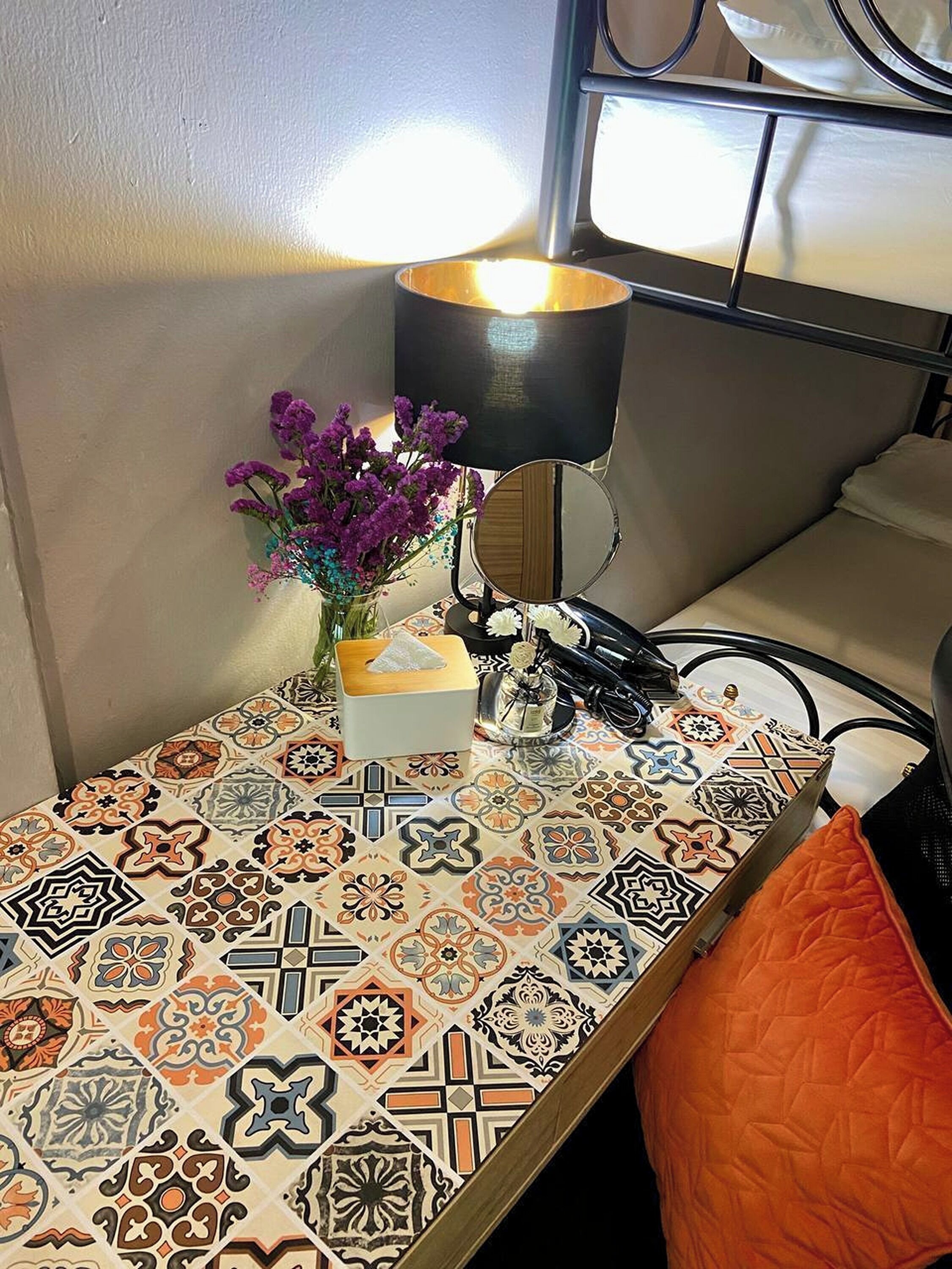A neatly arranged bedside table with a patterned tablecloth, a vase with purple flowers, a tissue box, a small bottle, and a lamp.