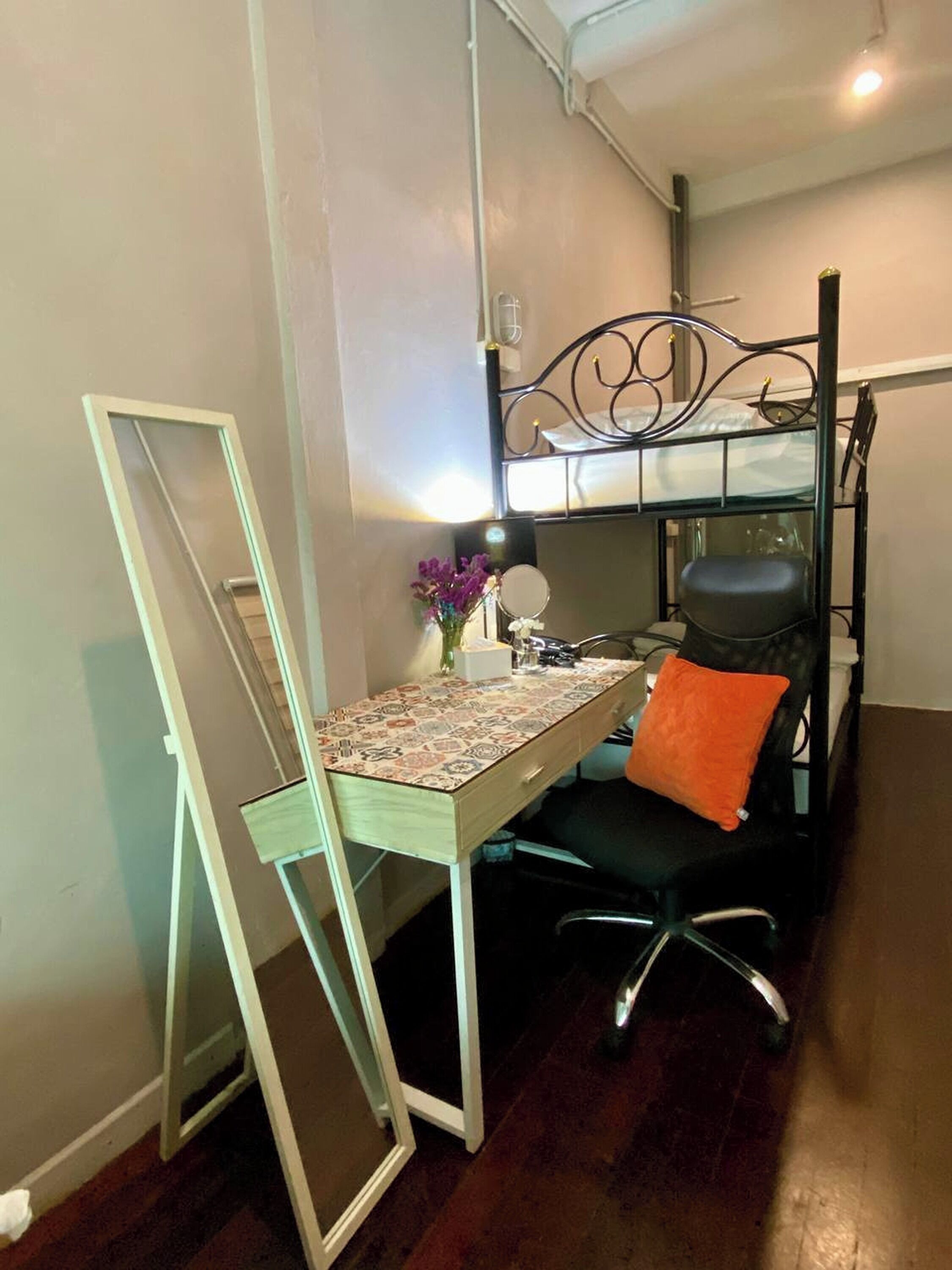 A compact room with a bunk bed, a desk with a chair, a mirror, and a small table with a vase of flowers.