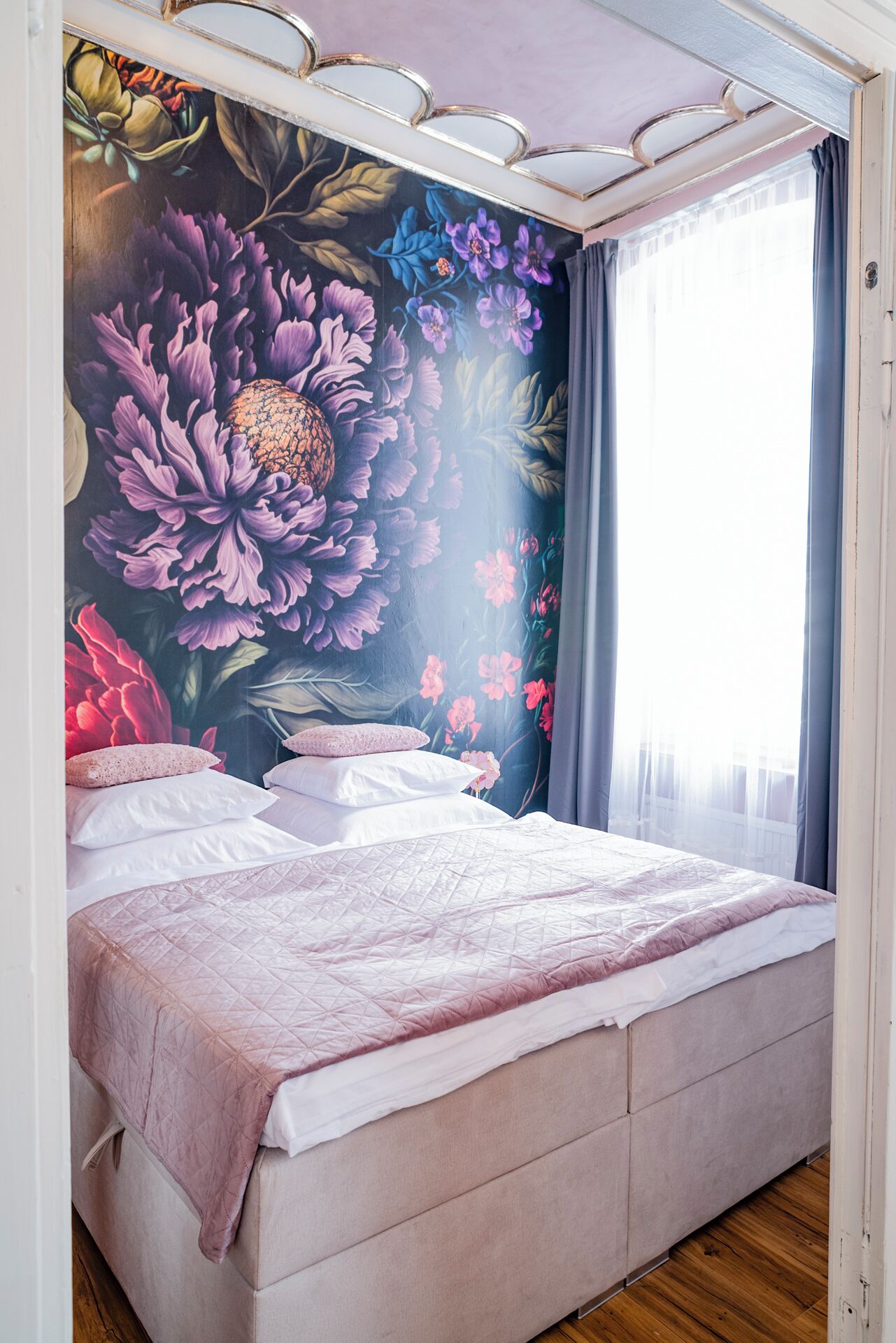 Un dormitorio con un amplio mural floral en la pared, una cama con colcha rosa y una ventana con cortinas.