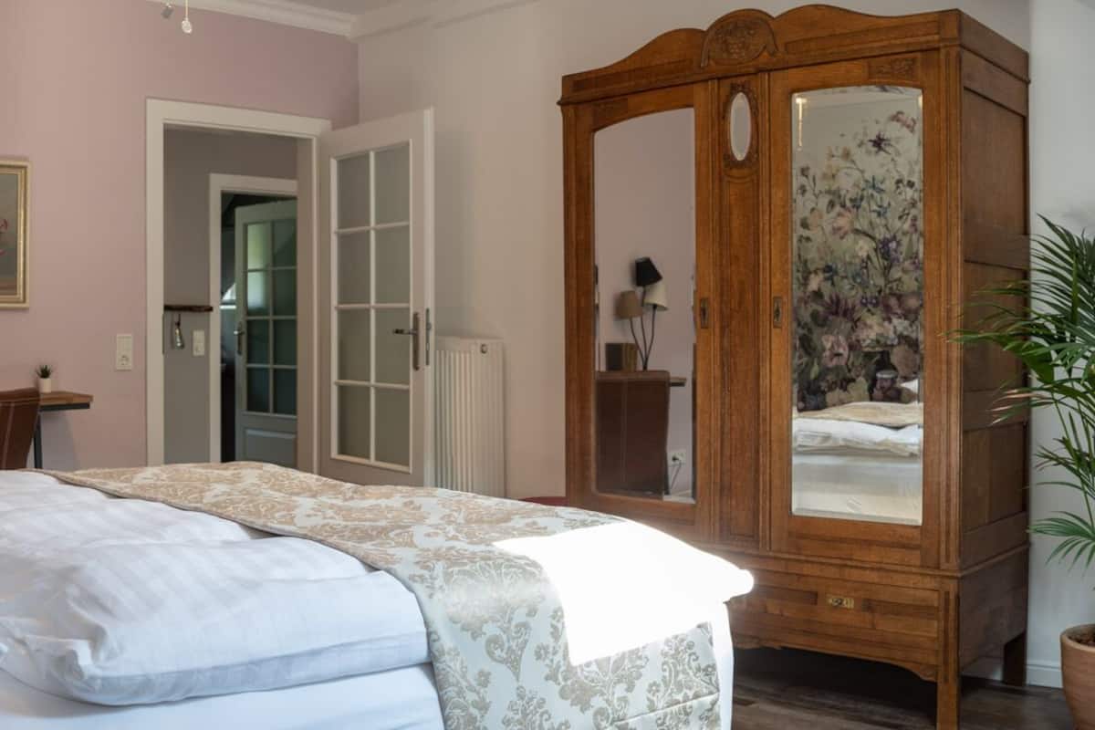 A bedroom with a bed, bedside lamps, a wooden wardrobe, and a door leading to another room.