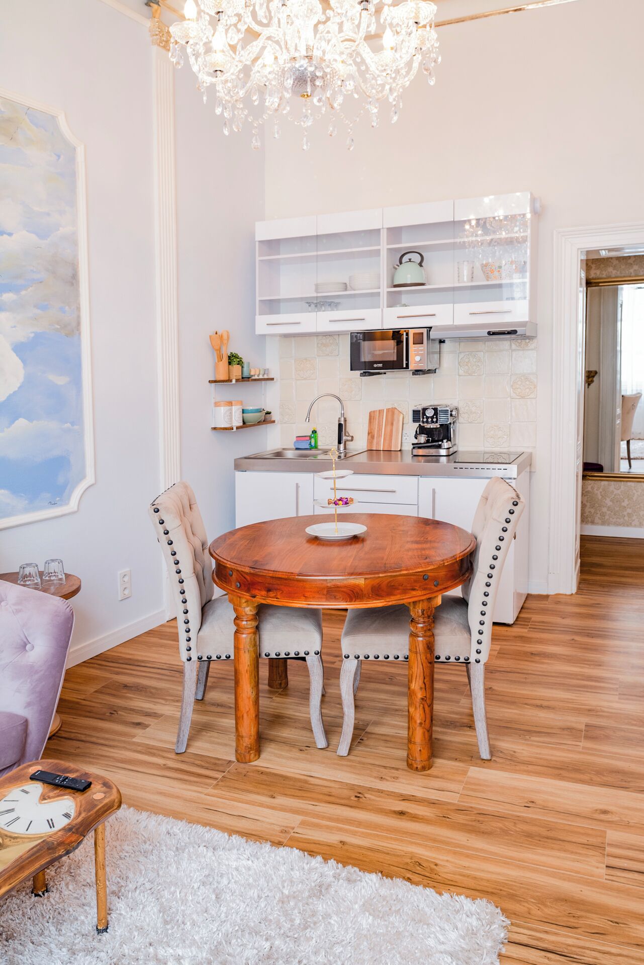 A dining area with a round wooden table, two upholstered chairs, a chandelier, and a kitchen in the background.