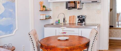 A dining area with a round wooden table, two upholstered chairs, a chandelier, and a kitchen in the background.