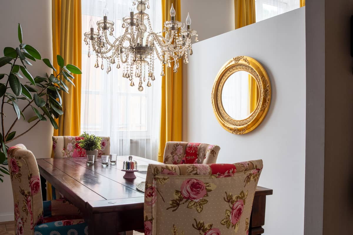A dining area with a wooden table, floral upholstered chairs, a chandelier, and a gold-framed mirror on the wall.