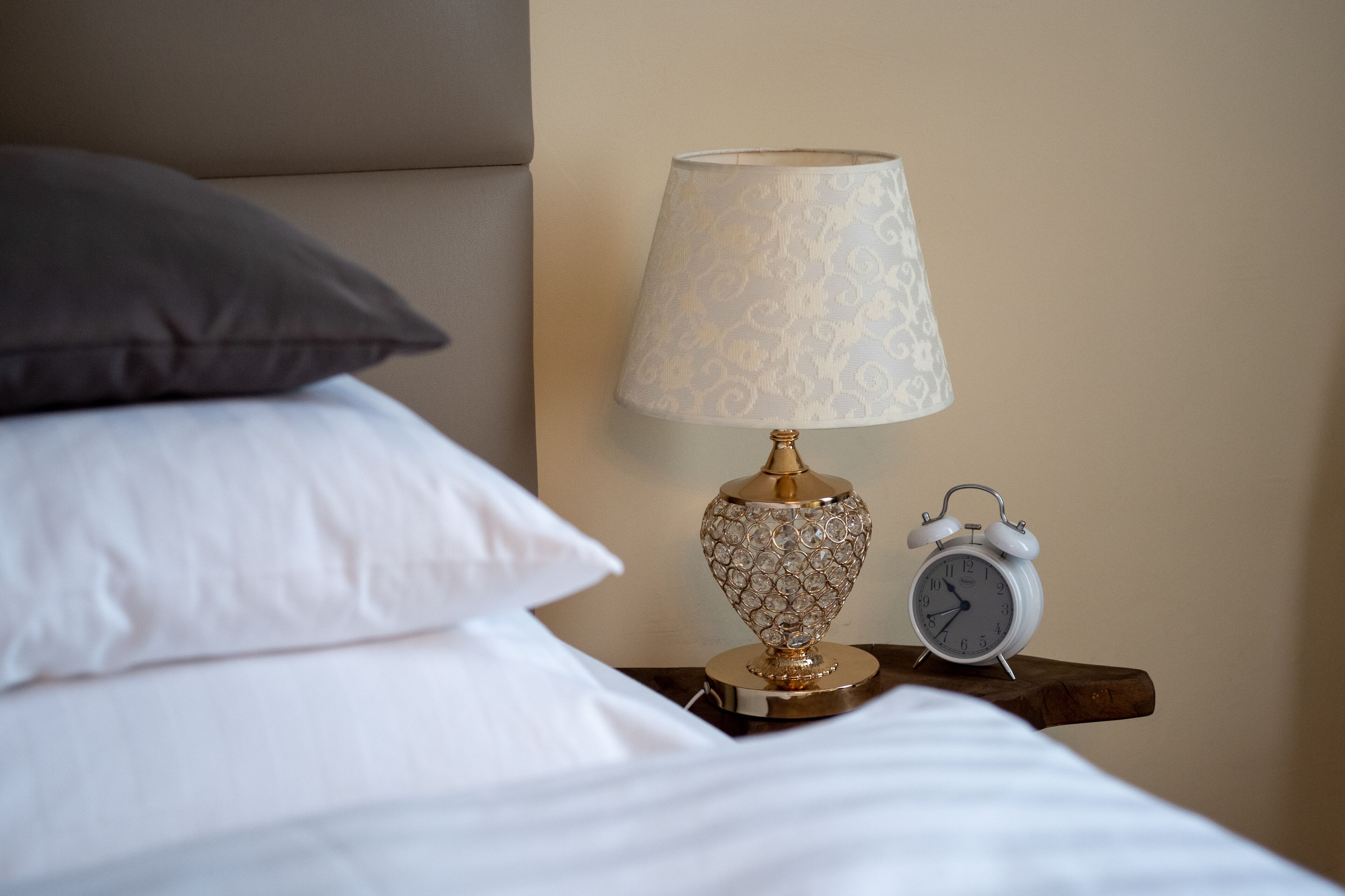 A neatly made bed with white linens and a dark pillow, a decorative lamp with a patterned shade, and a classic alarm clock on a wooden nightstand.