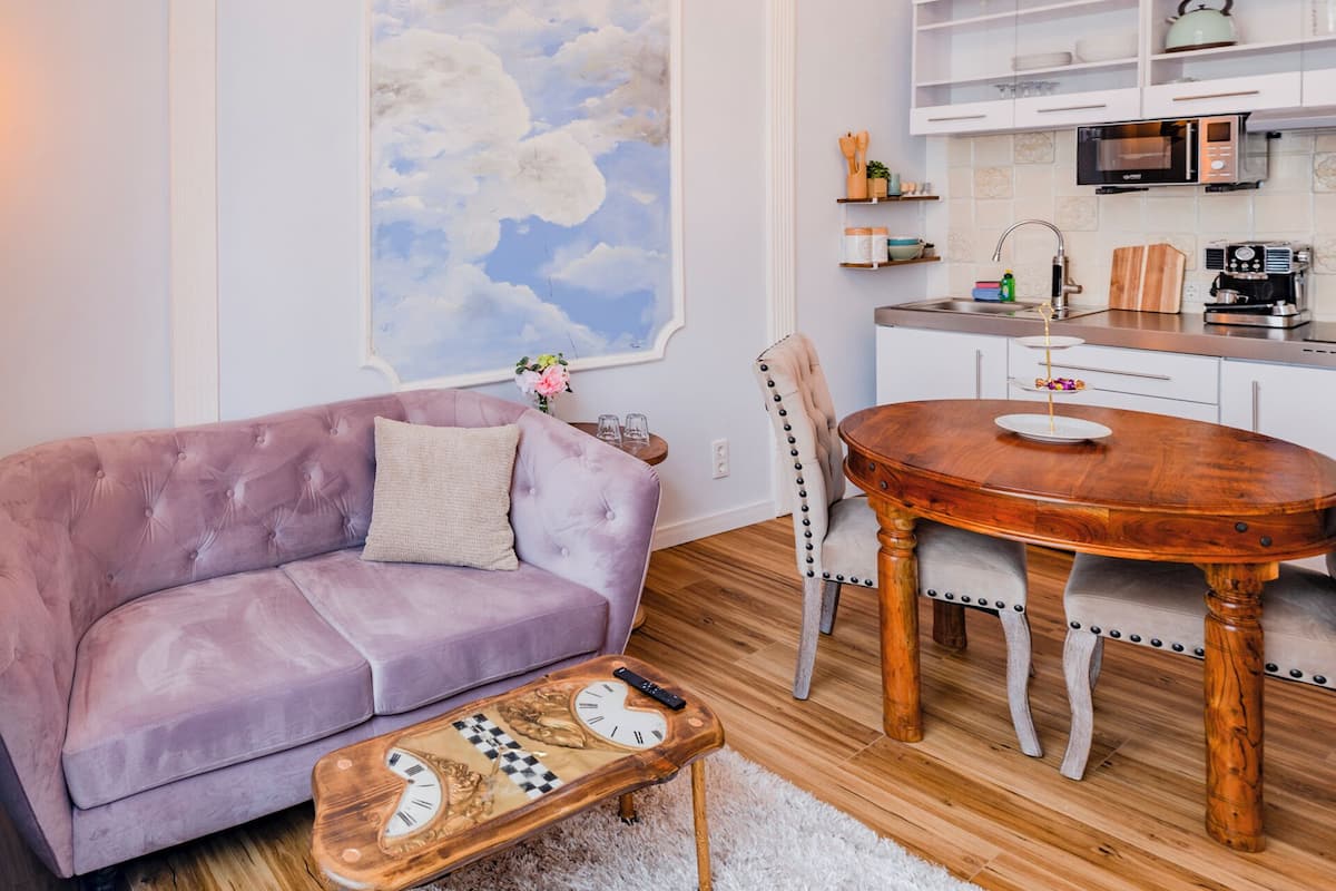 A living room with a purple sofa, a wooden coffee table with a clock, a dining table with chairs, a kitchen with white cabinets, and a large cloud painting on the wall.