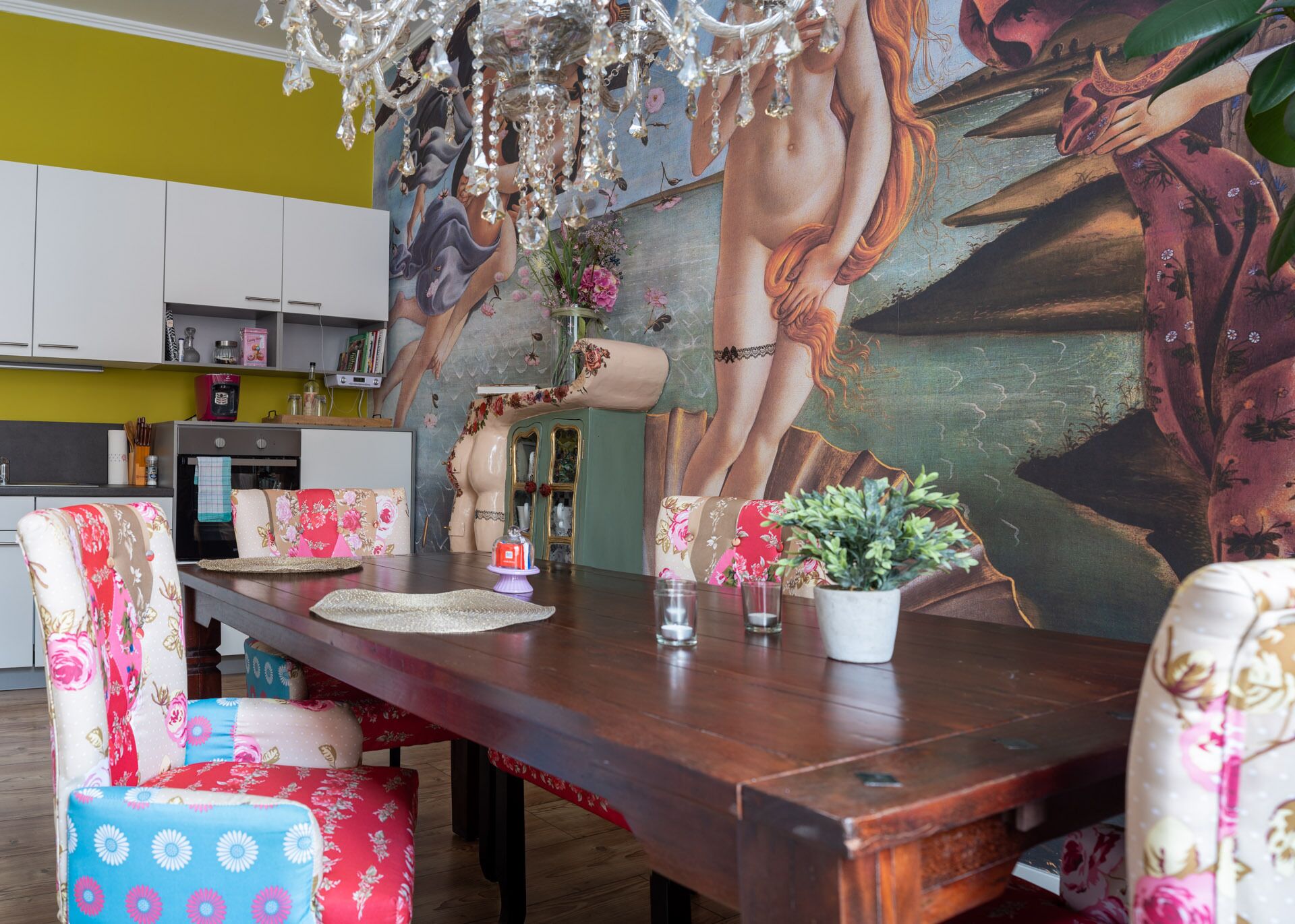 Un coin repas avec une table en bois, des chaises à motifs floraux, un lustre et une fresque murale.