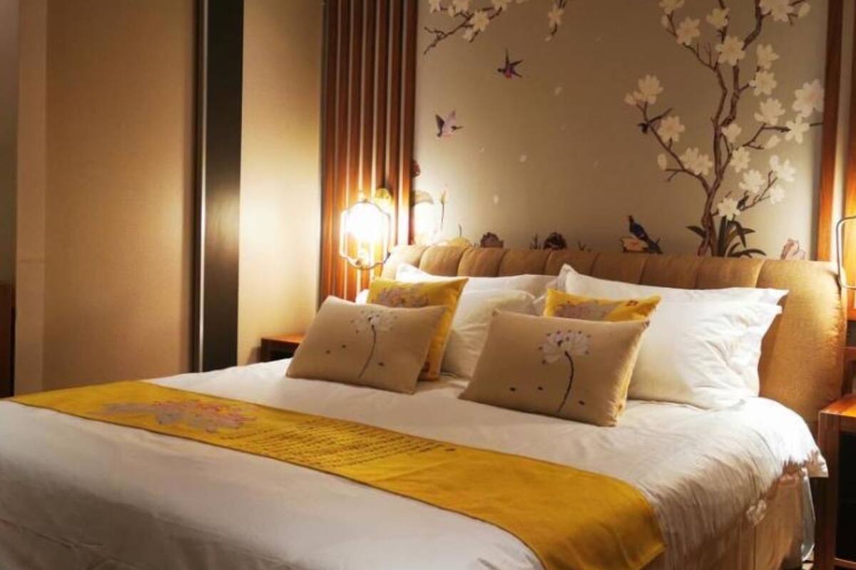 Un lit bien fait, avec une literie blanche et jaune, un mur décoratif orné de motifs floraux et d’oiseaux, et une source de lumière douce et chaleureuse.