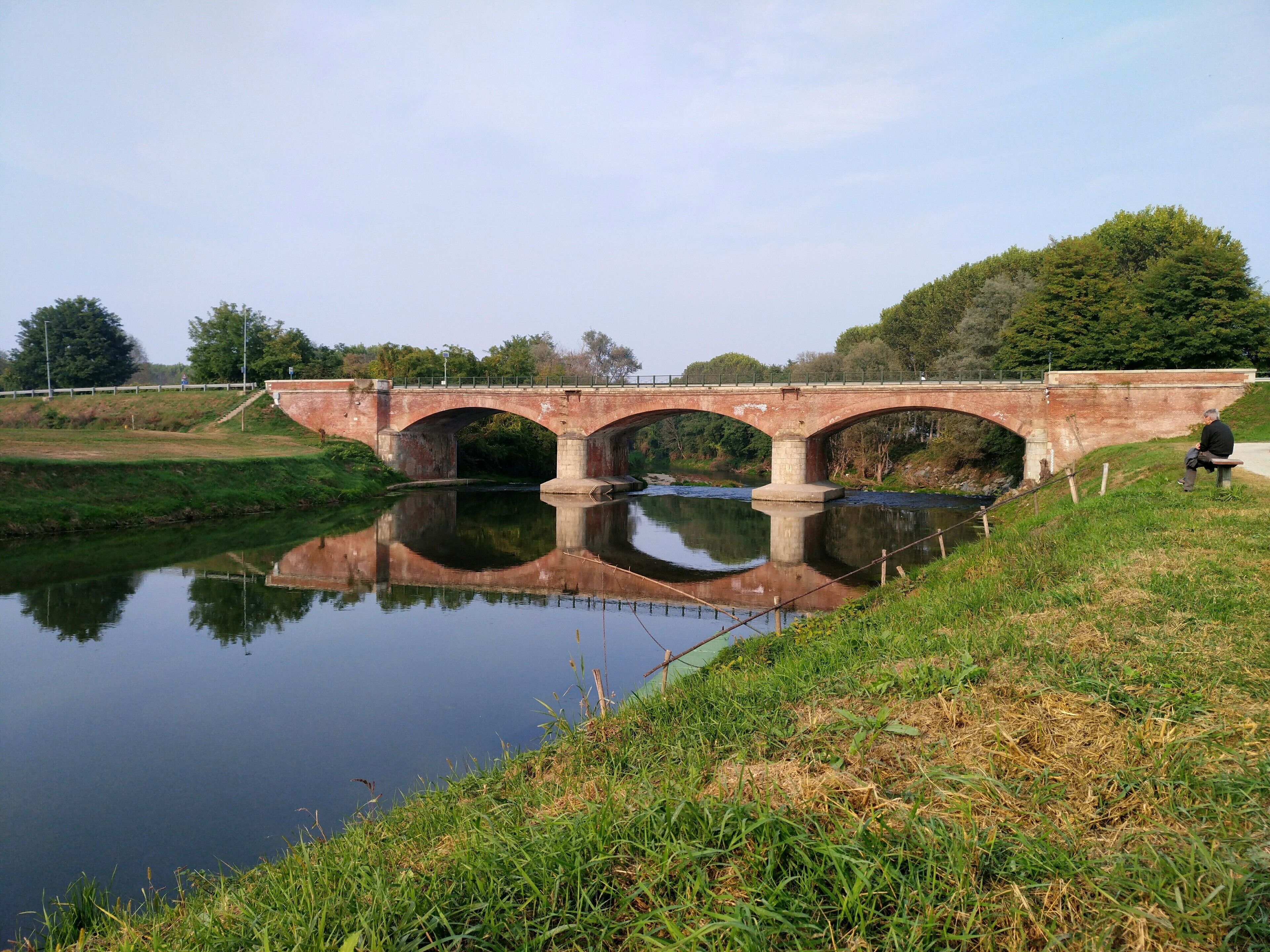 Eine Steinbrücke über einem ruhigen Fluss mit einer grünen Uferbank und Bäumen im Hintergrund.
