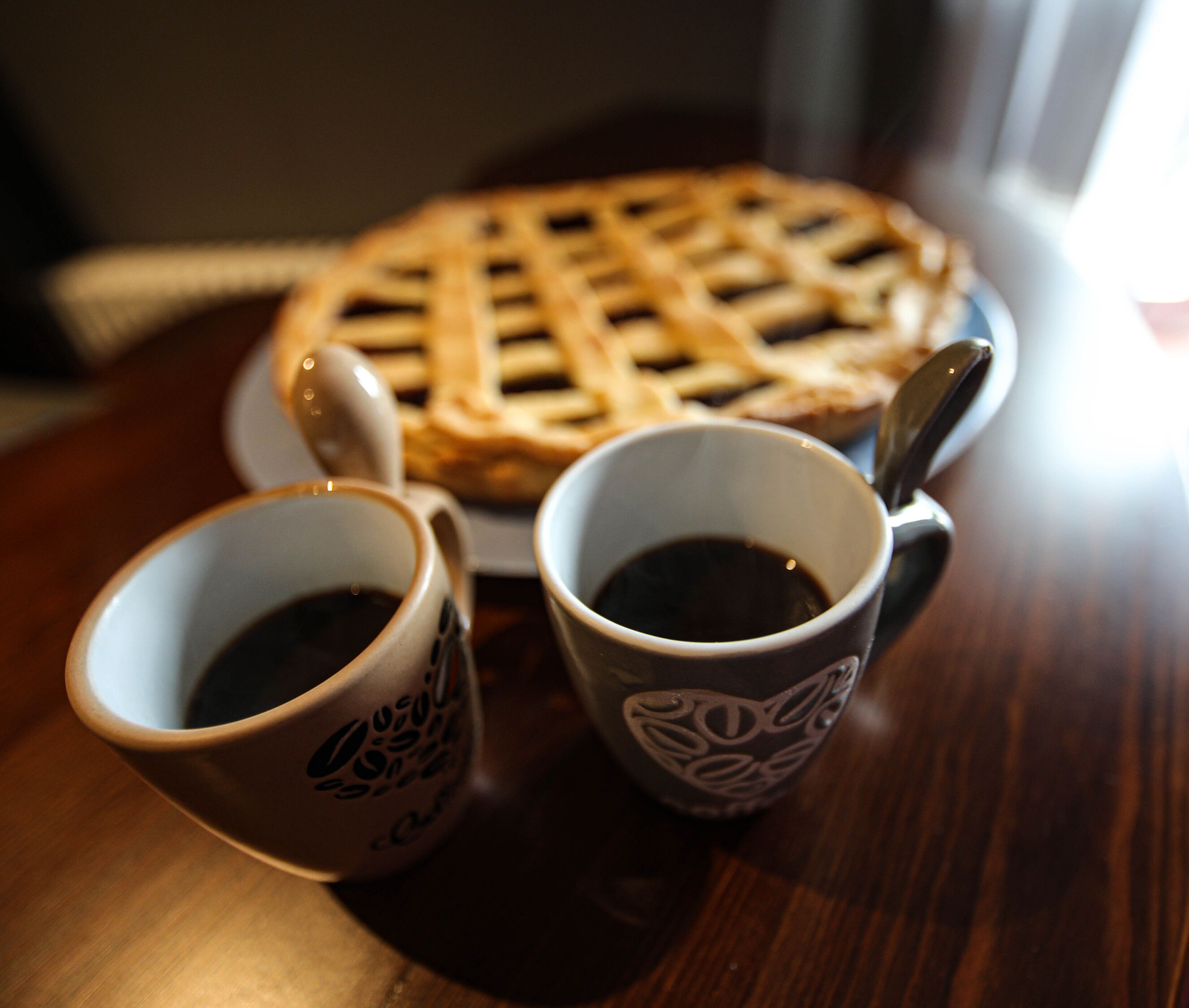 A close-up of a lattice-topped pie on a plate, accompanied by two cups of coffee on a wooden table.