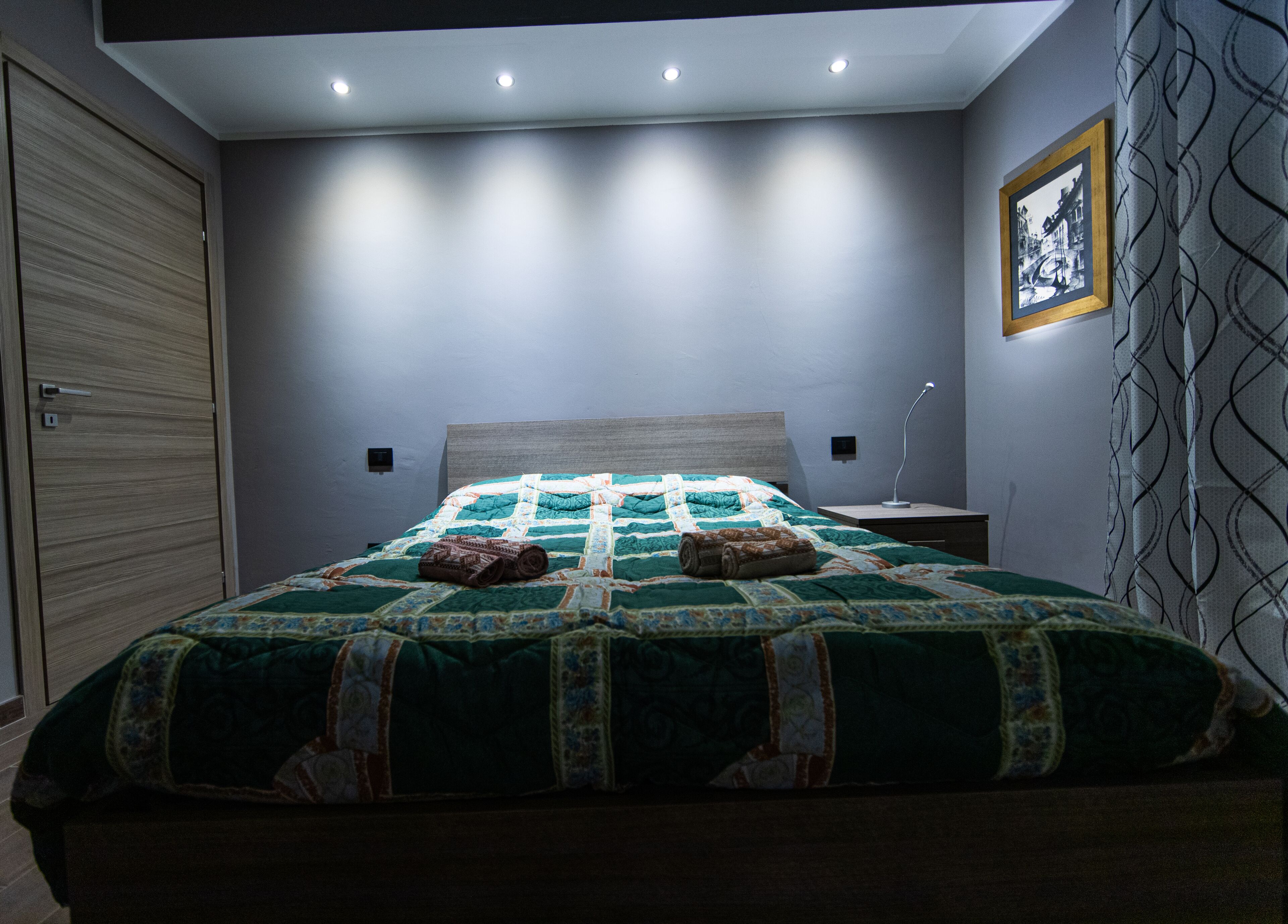 A bedroom with a bed, a nightstand, a lamp, a framed picture on the wall, and a patterned curtain.
